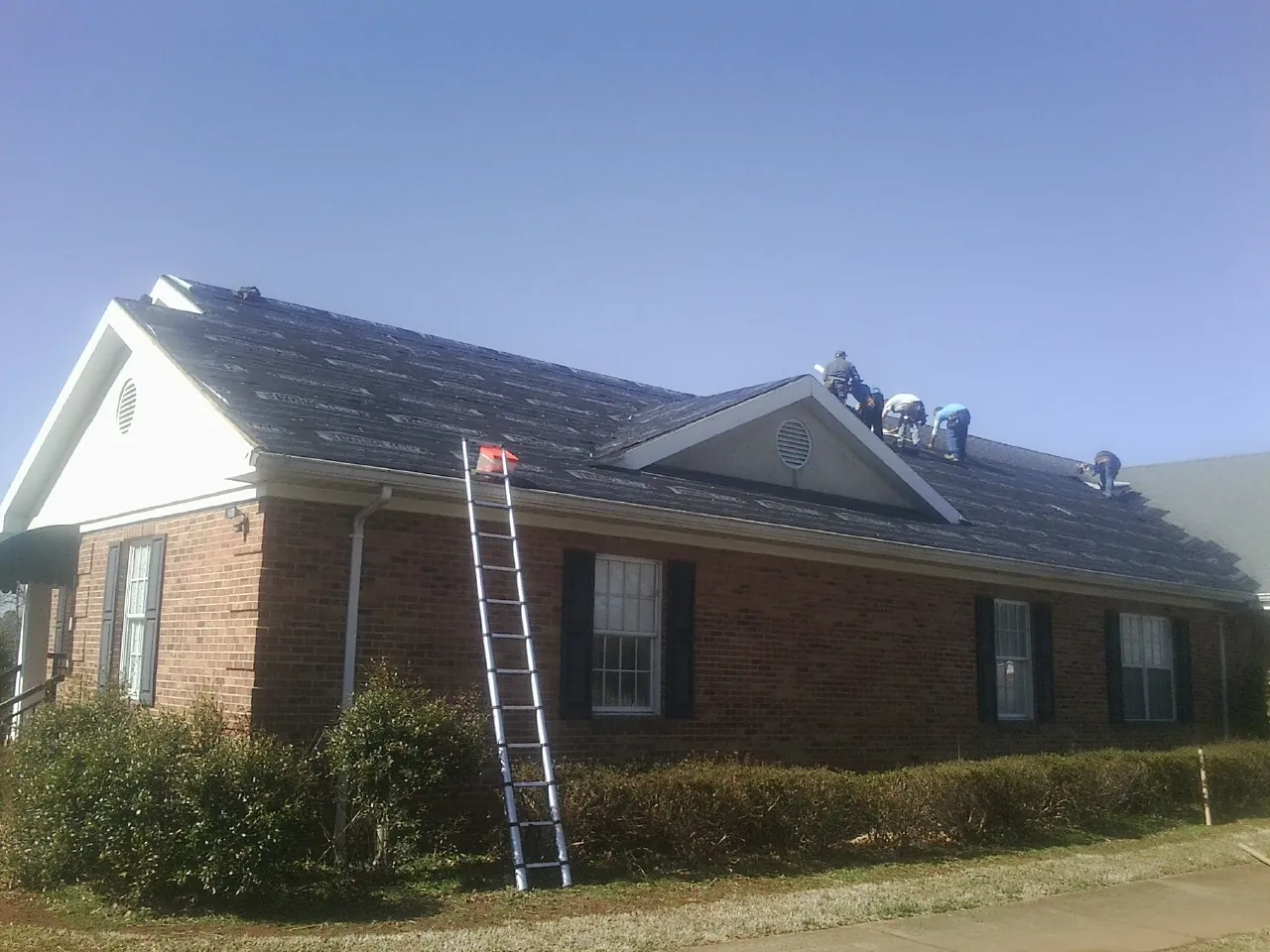 Crew tearing shingles off a brick ranch roof with a ladder set — Emergency Roof Repair work by our crew in Millbrook